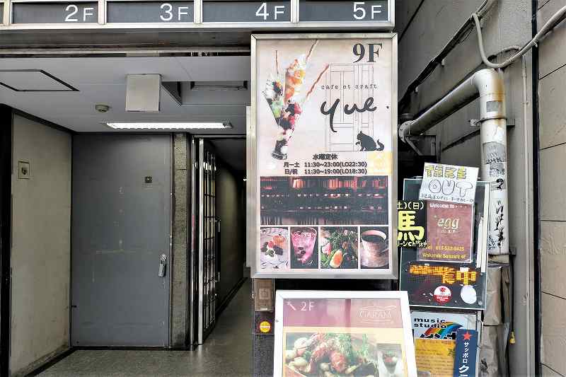 「カフェ エ クラフト ユエ」の案内看板がビルの1階に掲示されている